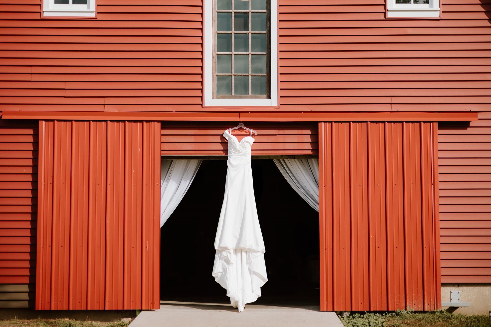 A wedding dress is hanging in the doorway of a red barn.