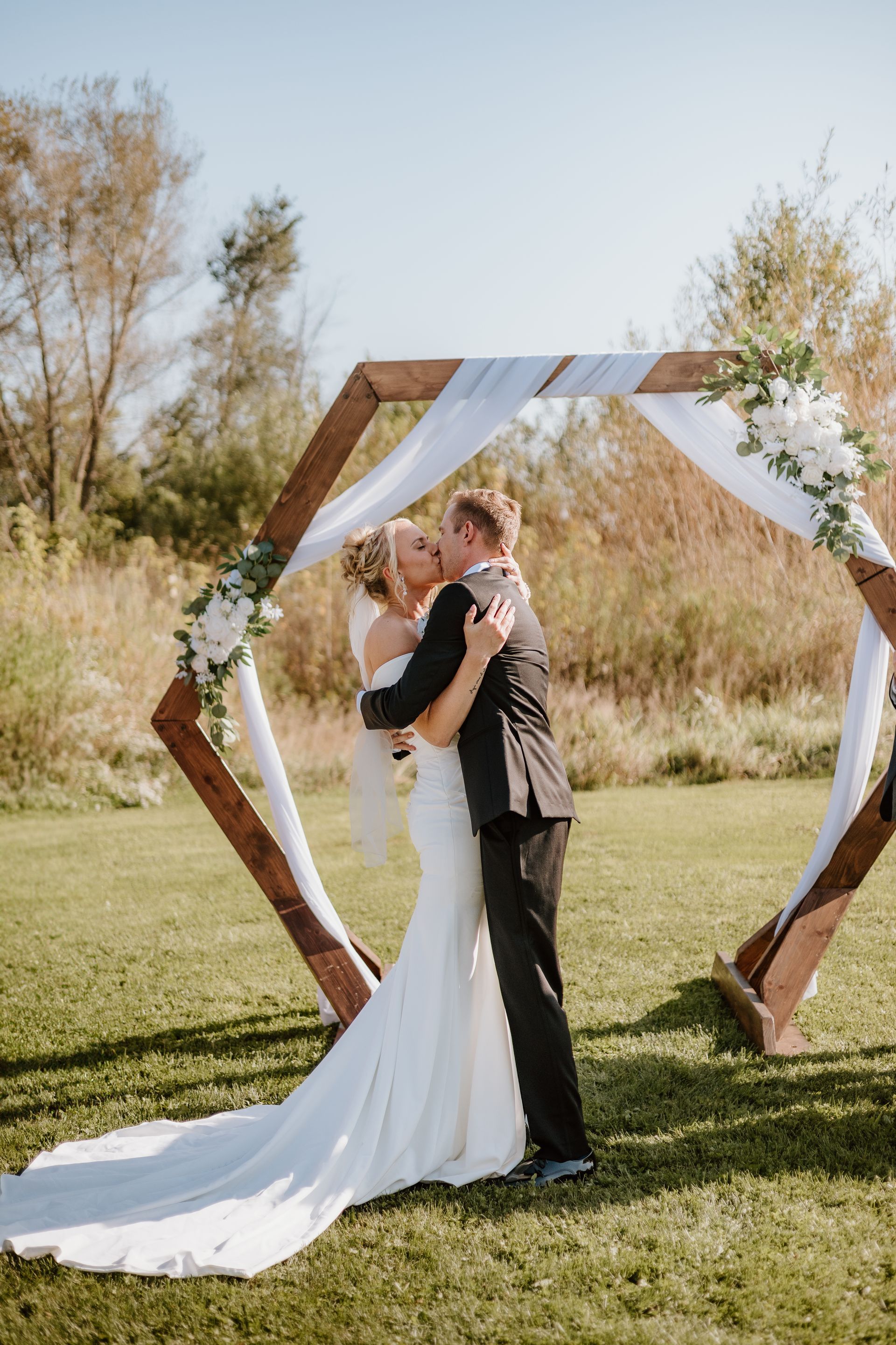 A bride and groom are kissing under a wooden arch at their wedding.