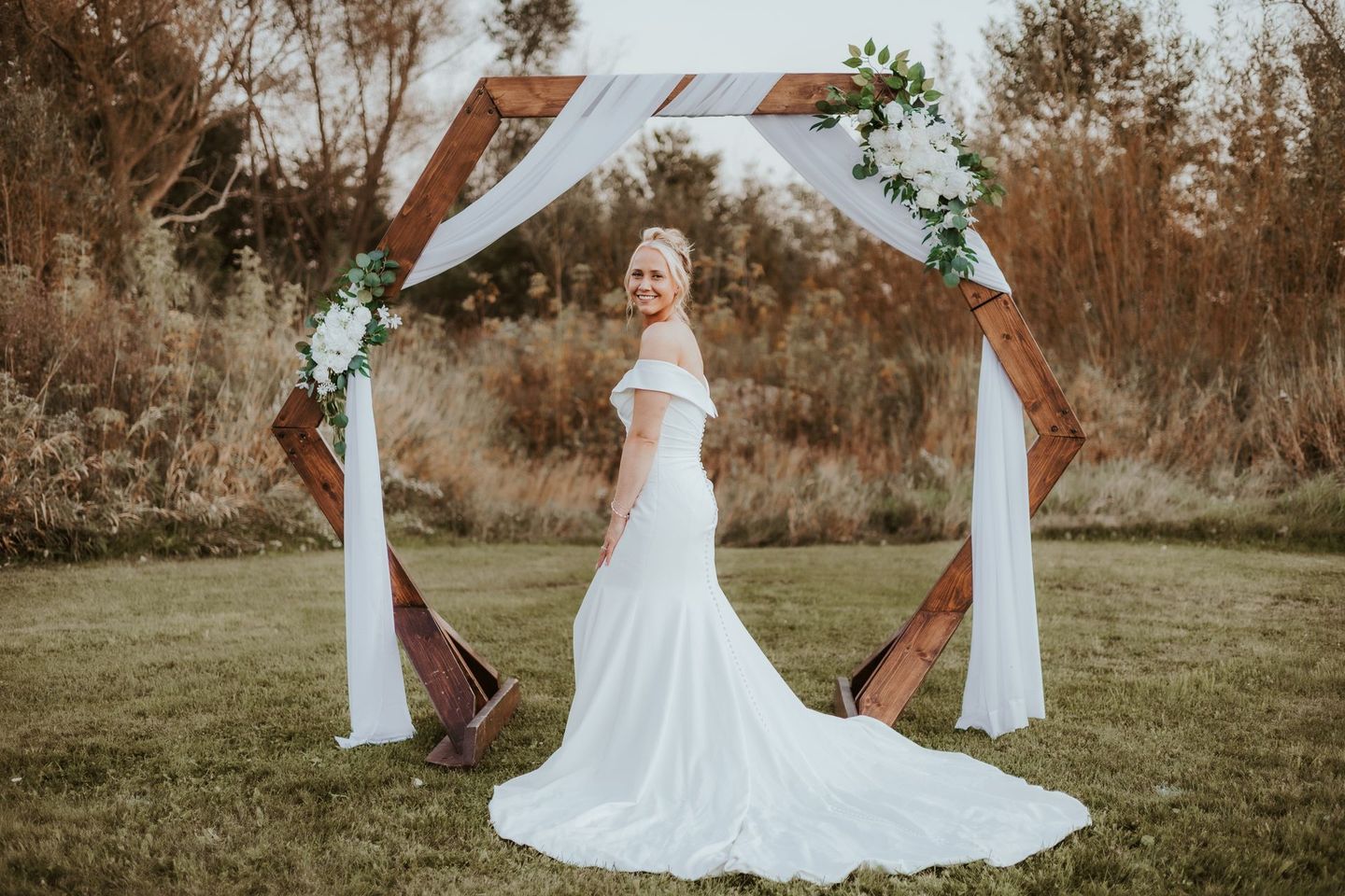 A bride in a wedding dress is standing in front of a wooden arch.