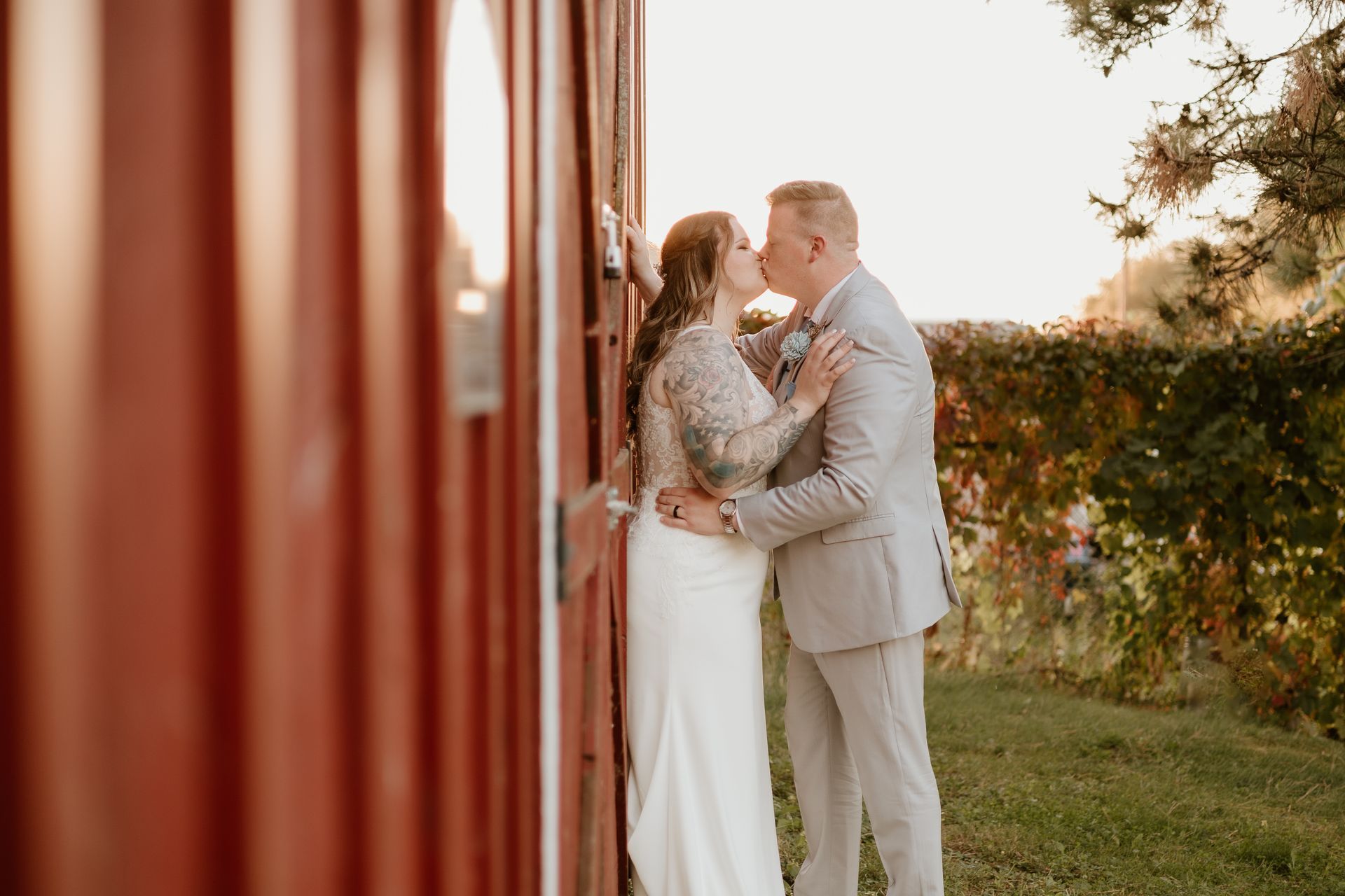 A bride and groom are kissing in front of a red barn.