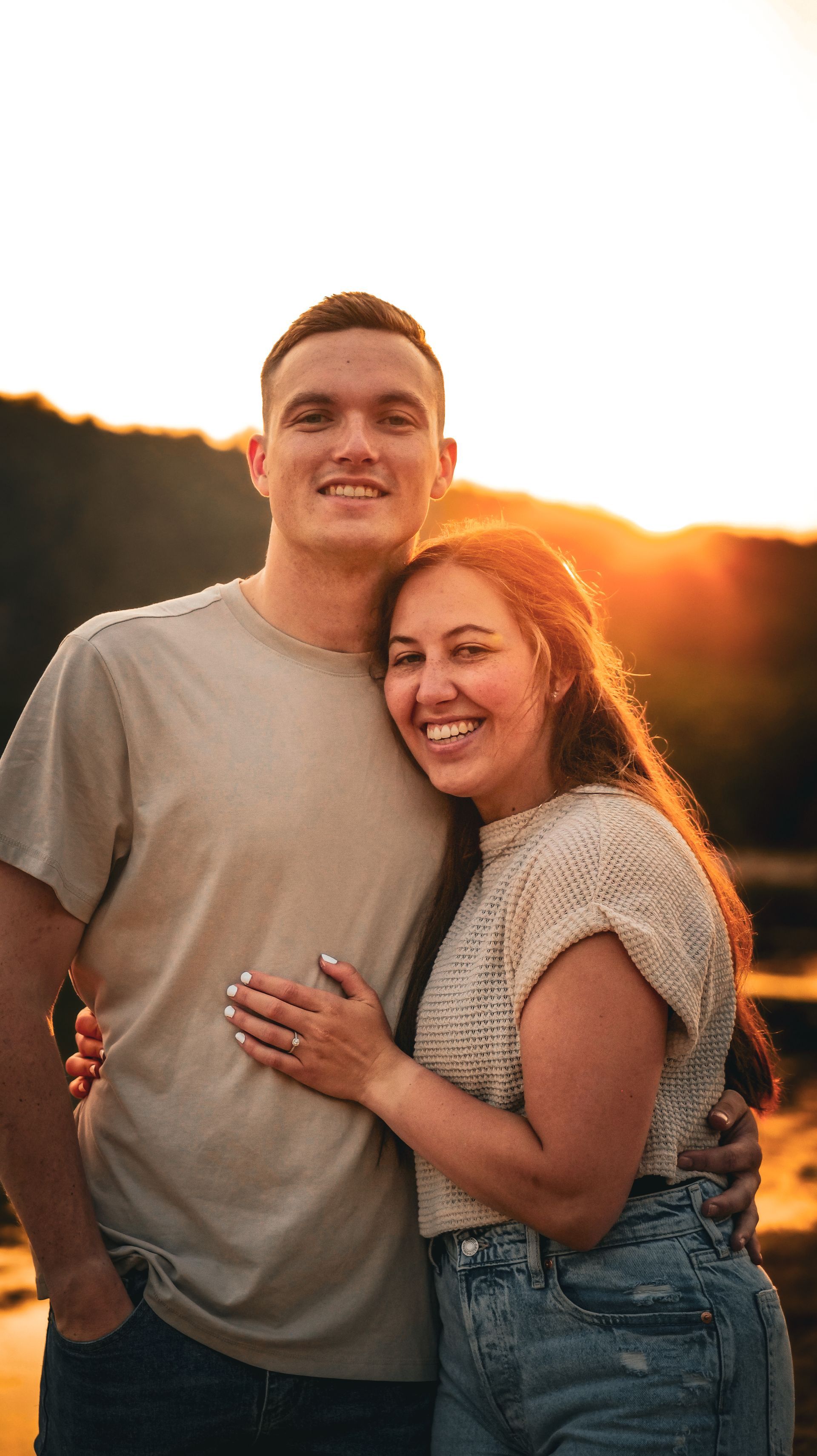 A man and a woman are posing for a picture in front of a lake at sunset.