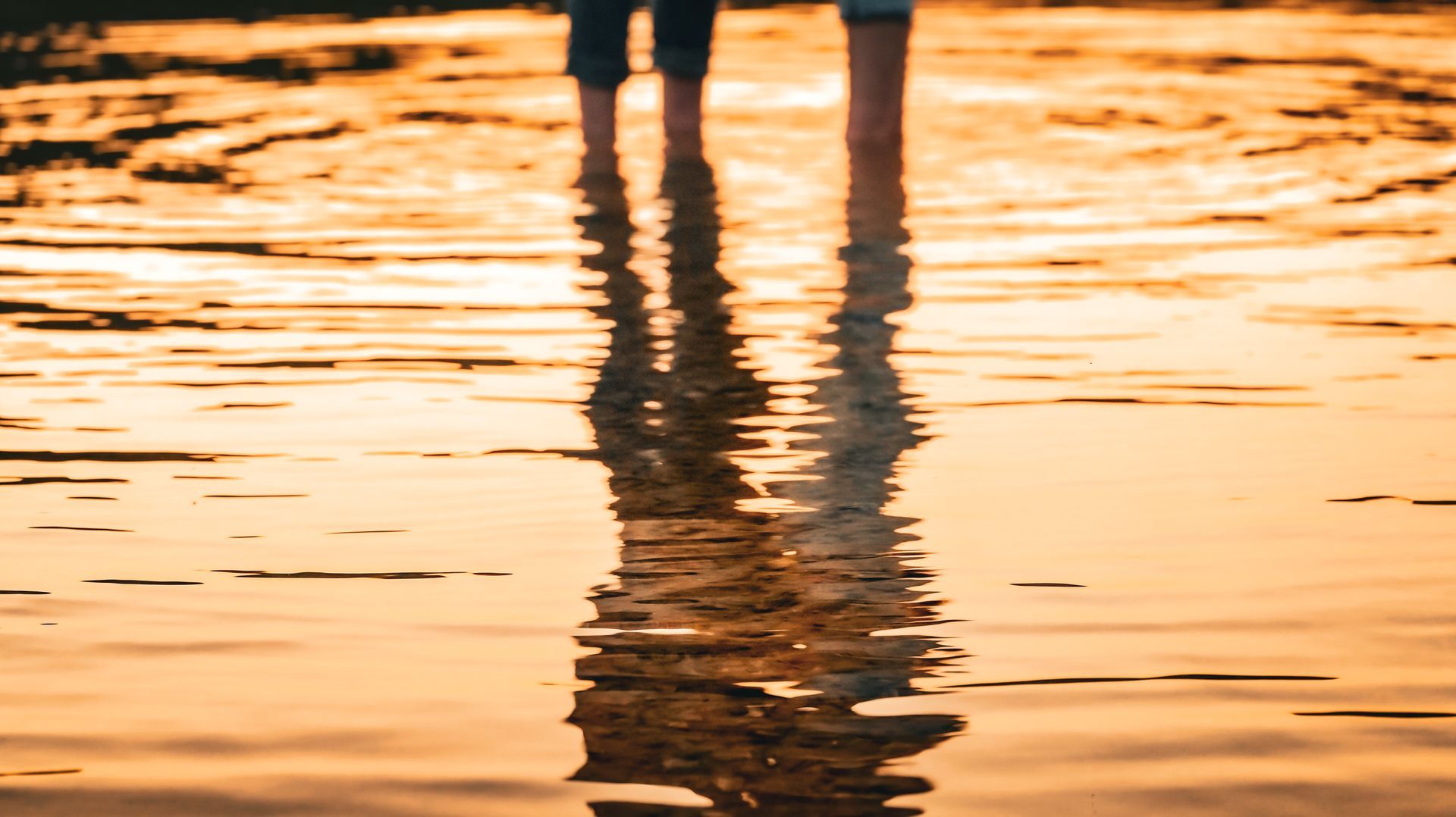A couple of people standing in the water on a beach at sunset.