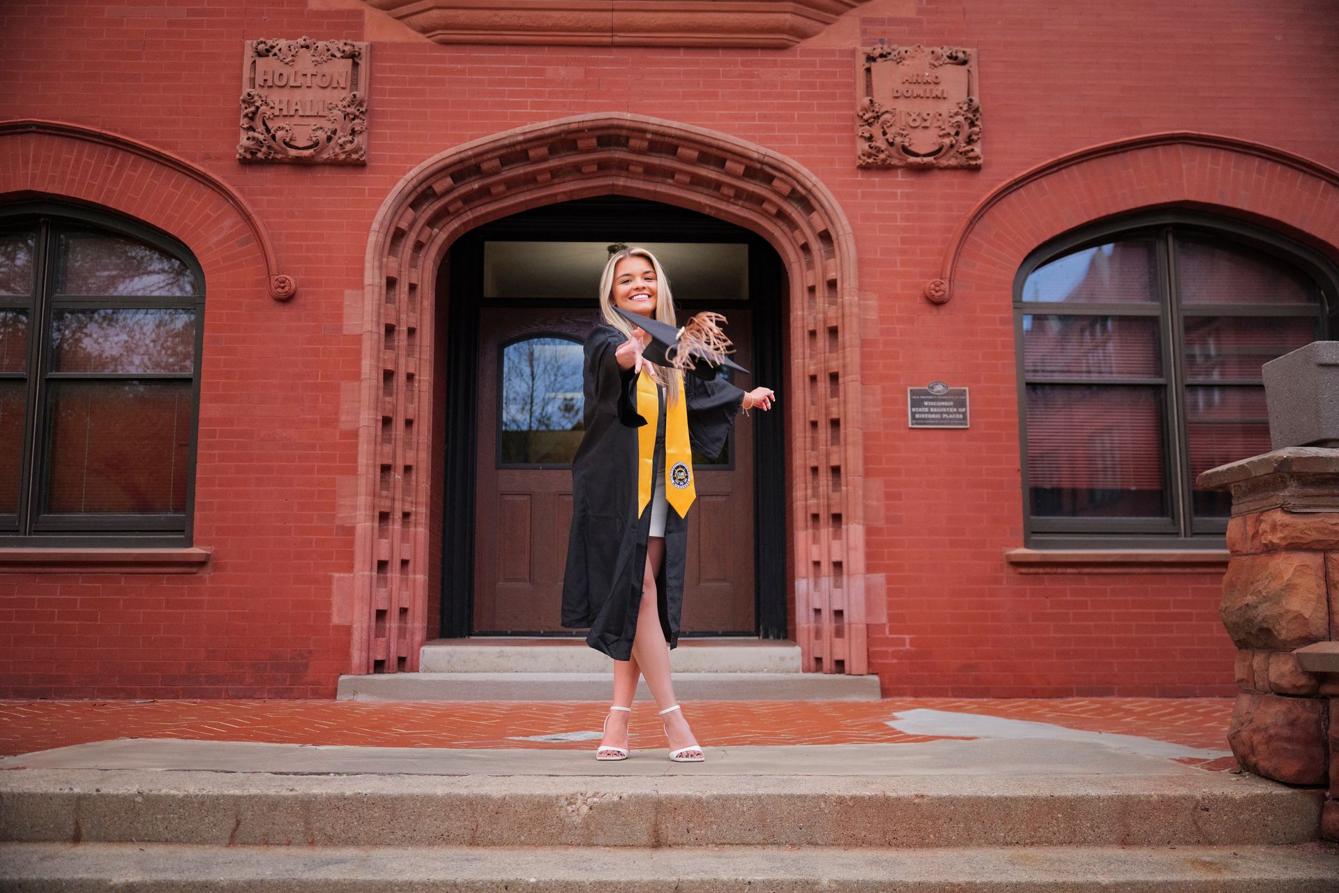 A woman in a graduation cap and gown is standing in front of a brick building.