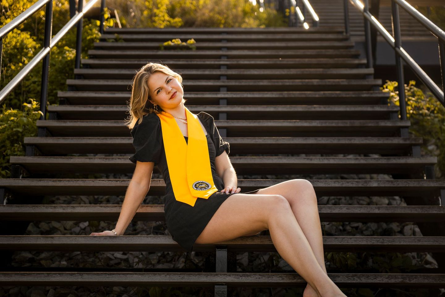 A woman in a black dress and yellow sash is sitting on a set of stairs.