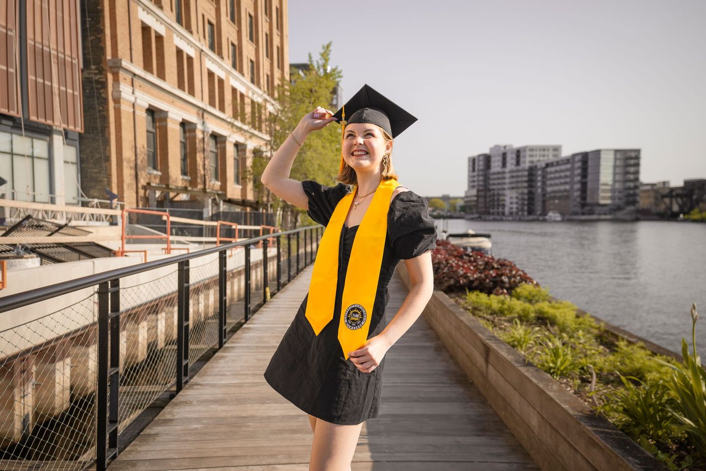 A woman in a graduation cap and gown is standing on a wooden walkway next to a body of water.