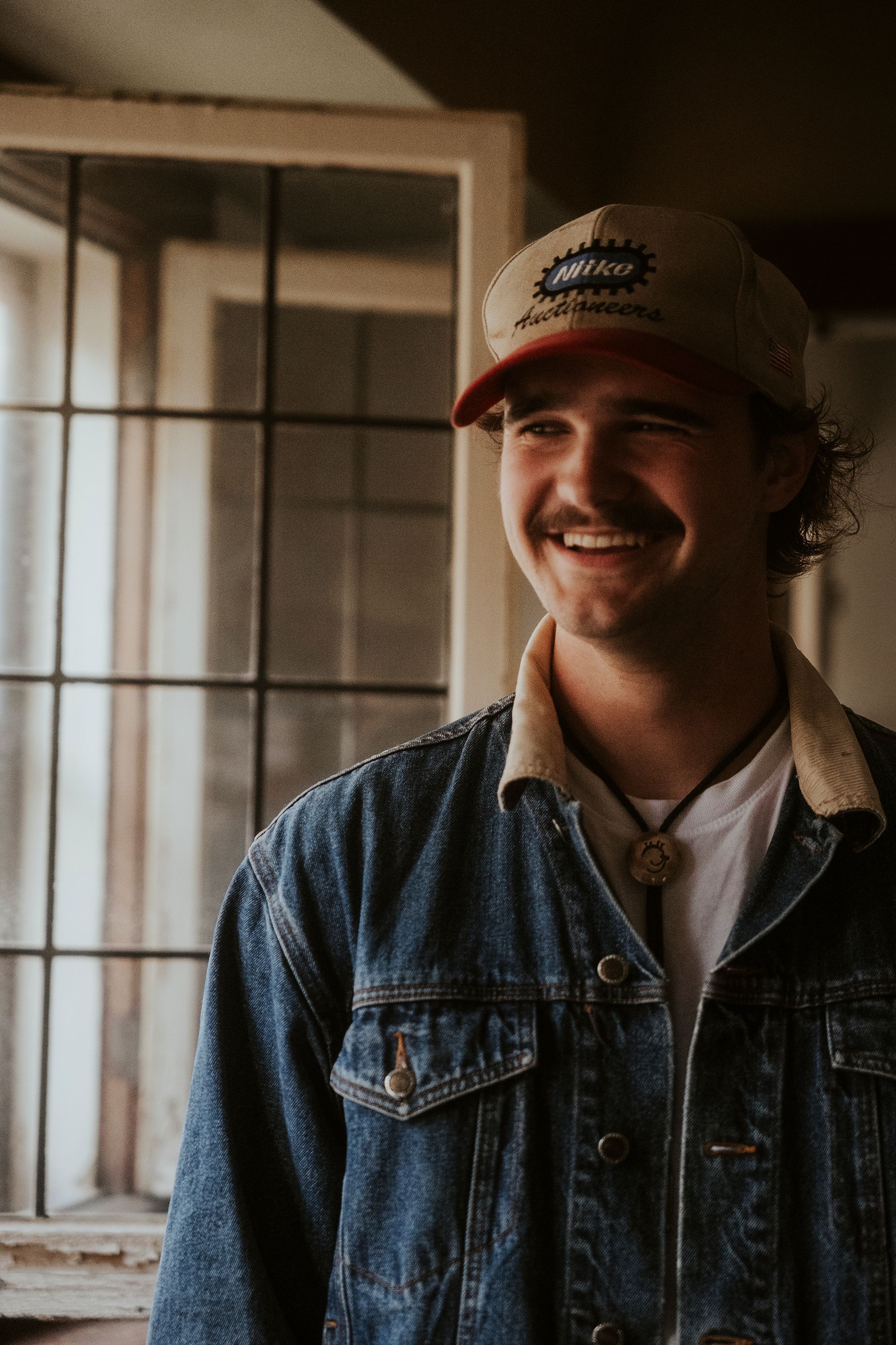 A man wearing a hat and a denim jacket is smiling in front of a window.