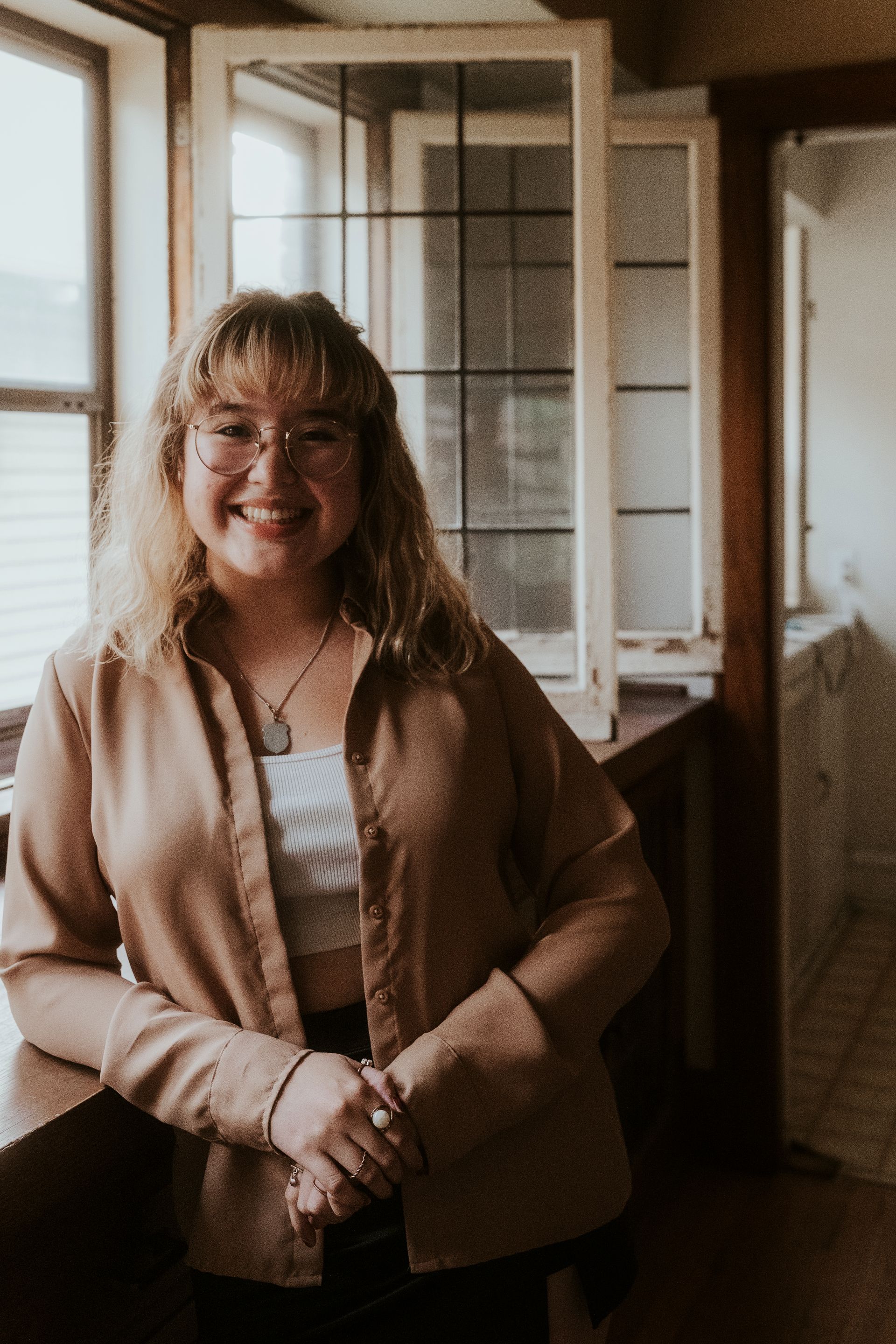 A woman wearing glasses and a brown shirt is standing in front of a window.