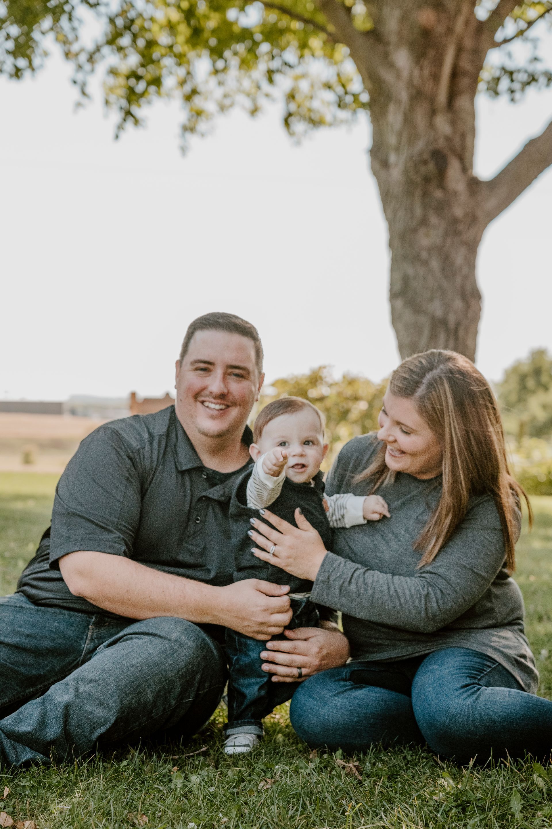 A family is sitting in the grass under a tree holding a baby.
