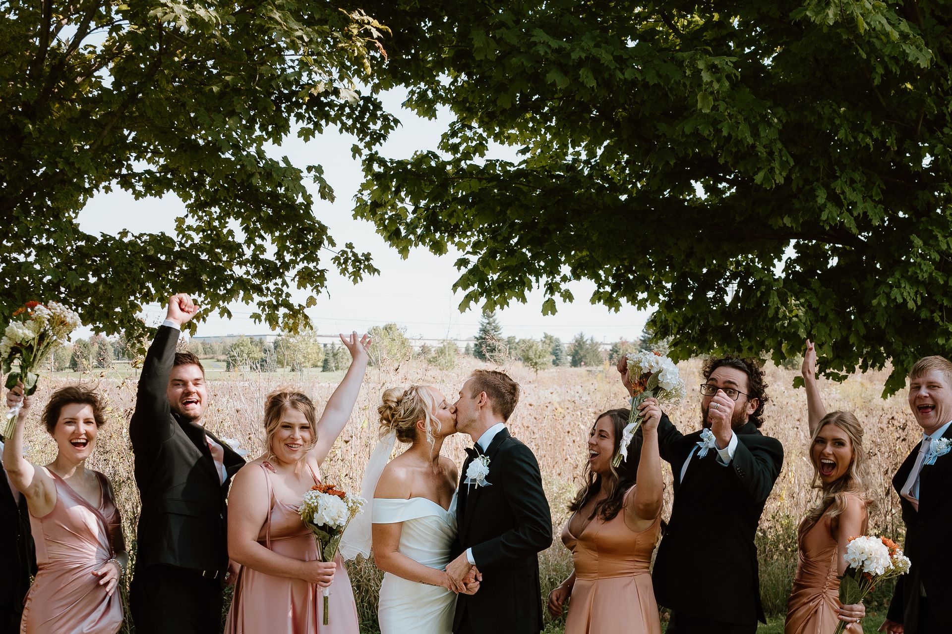 A bride and groom are kissing under a tree with their wedding party.