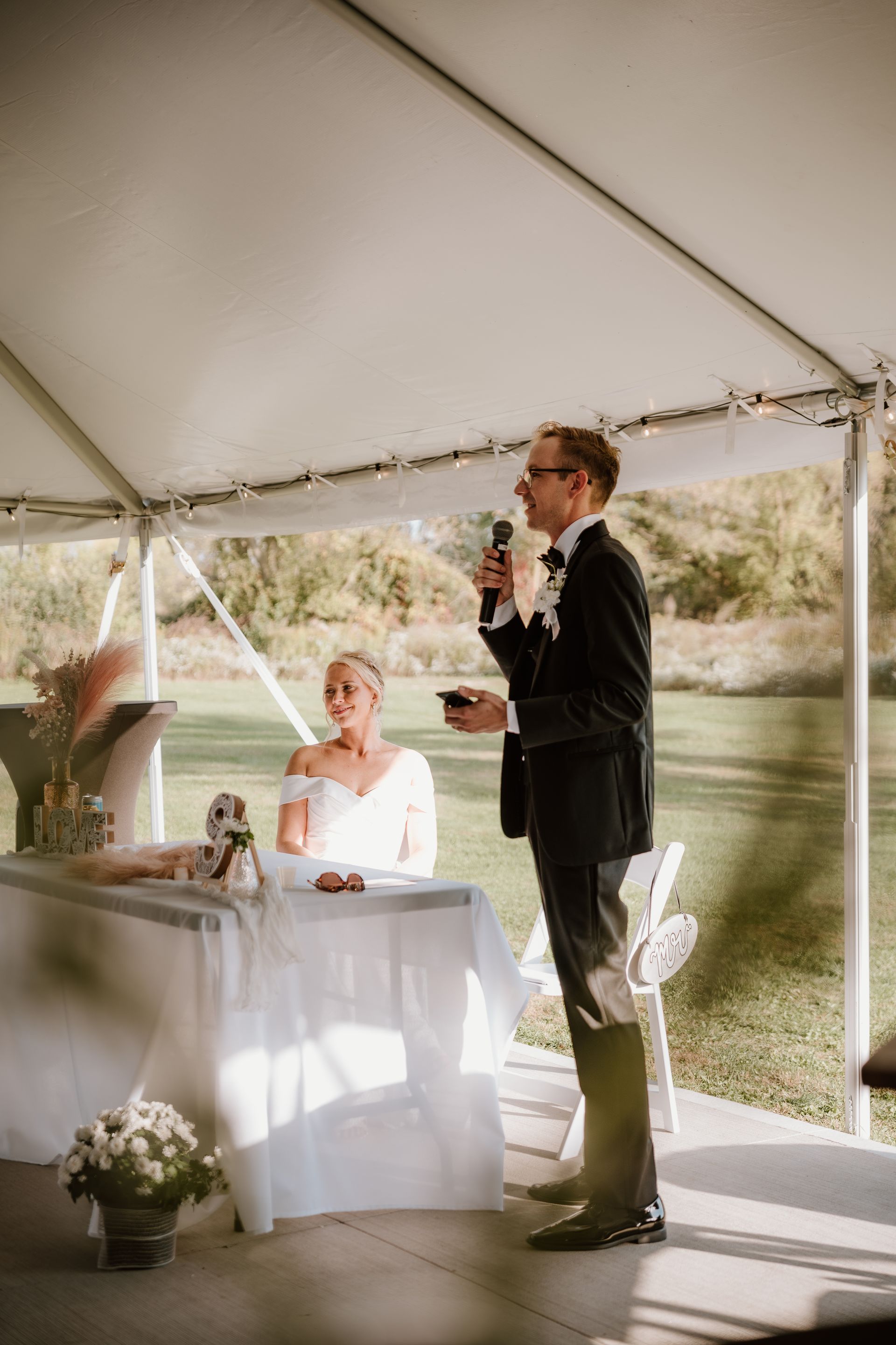 A bride and groom are sitting at a table under a tent while the groom is giving a speech.