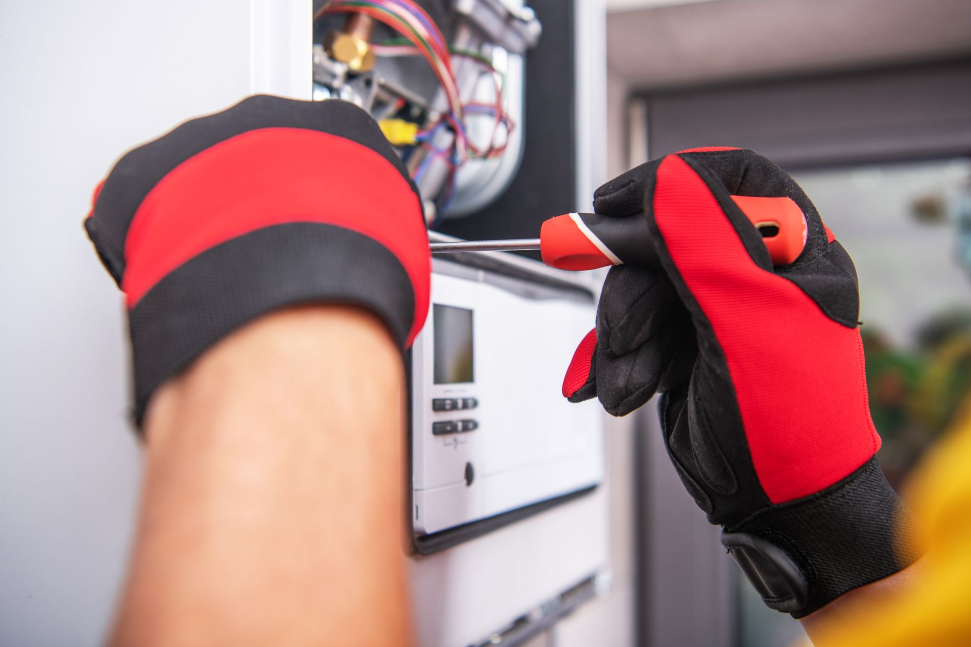 Hands wearing red and black gloves using a screwdriver on a white appliance.