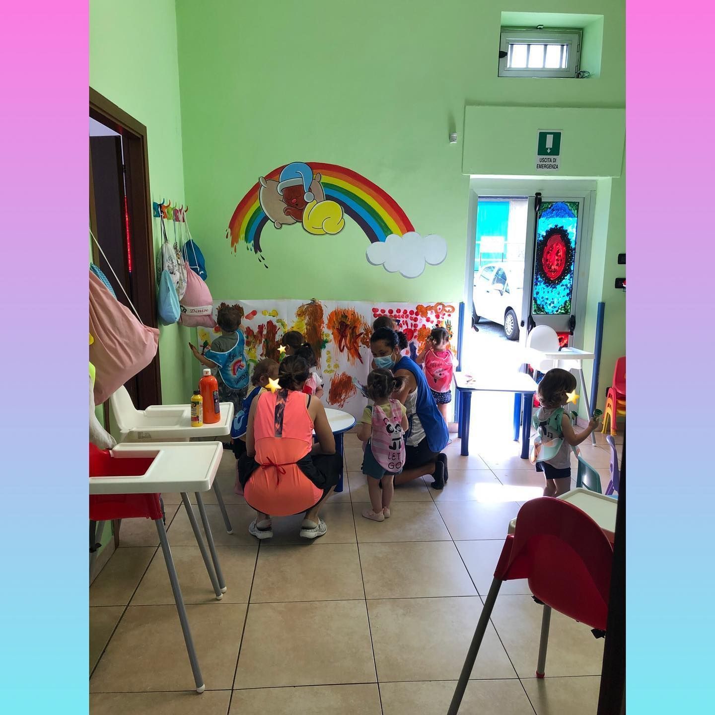 Bambini in una sala giochi con un'insegnante, che dipingono seduti a un tavolo. Un arcobaleno decora la parete e una porta conduce all'esterno.
