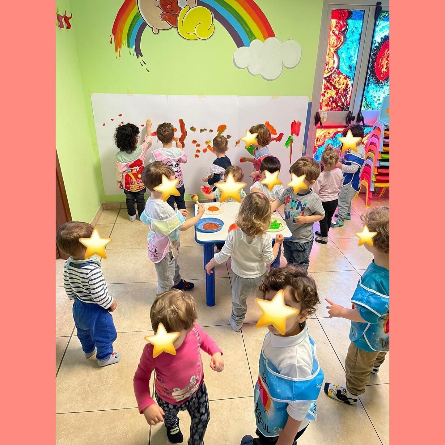 Bambini che dipingono a un tavolo e su una grande lavagna bianca in un'aula colorata. Un arcobaleno e delle nuvole decorano la parete.