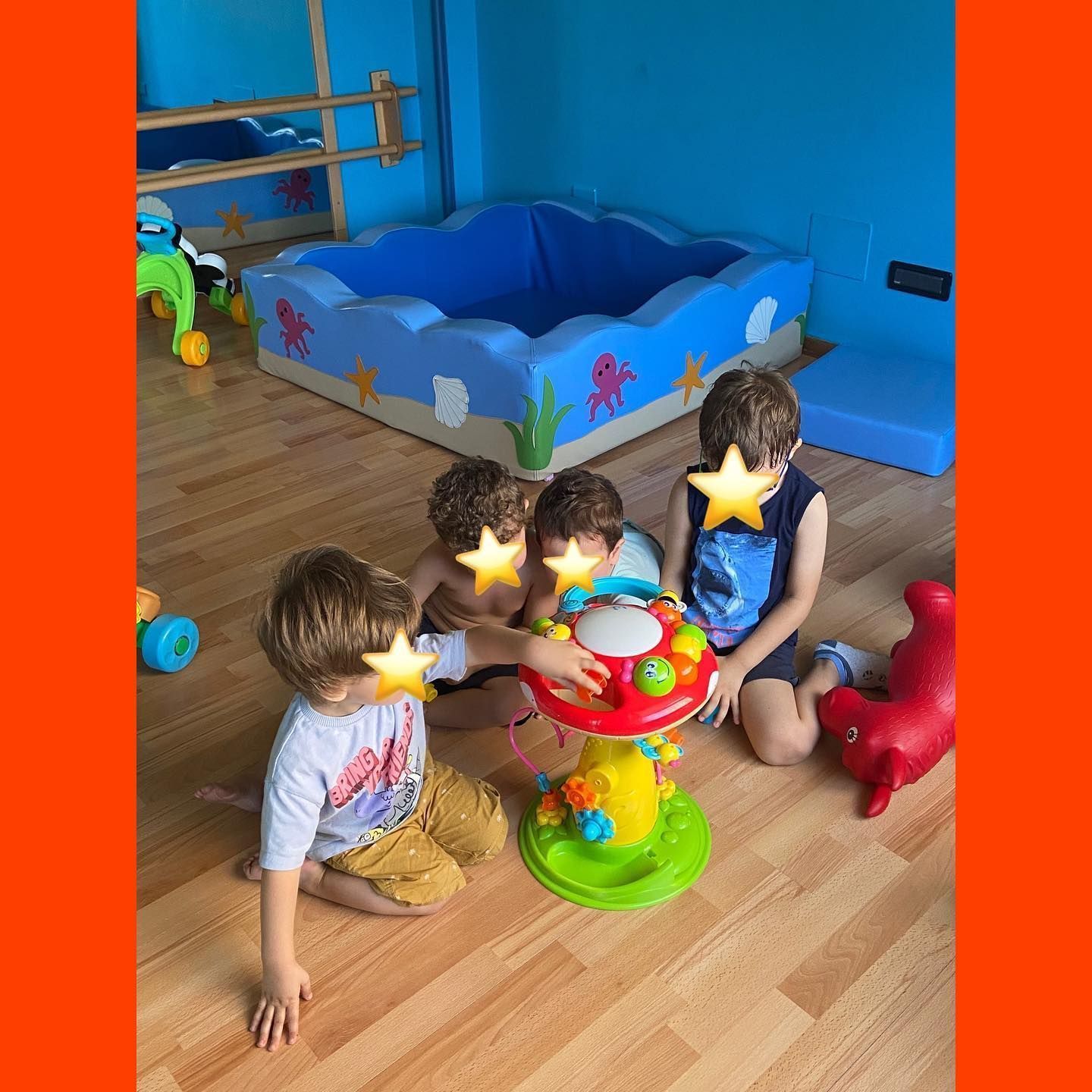 Quattro bambini giocano con un giocattolo su un pavimento di legno, vicino a una piscina di palline, in una stanza dai colori vivaci.