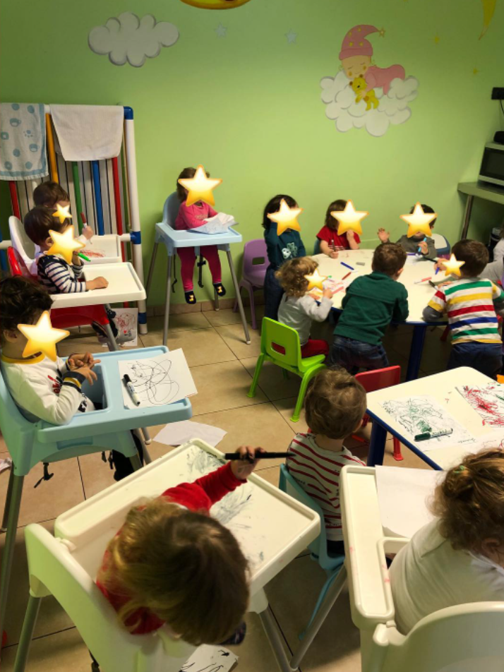 I bambini in classe disegnano seduti ai tavoli e sui seggioloni. La stanza è arredata con colori vivaci e ha una parete verde.