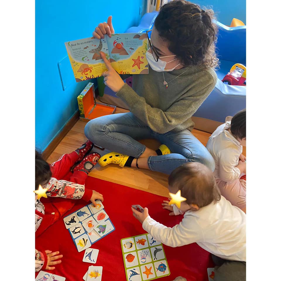 Un'insegnante con la mascherina legge un libro ai bambini seduti su un tappeto rosso, mentre altri giocano a un gioco di abbinamento in una stanza blu.