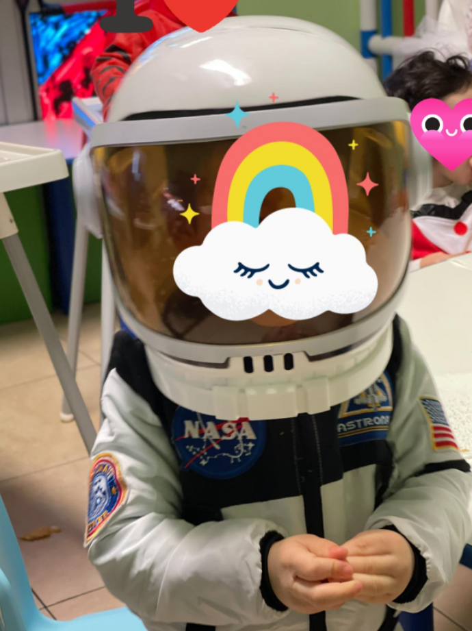 Bambino che indossa una tuta e un casco da astronauta della NASA, sorridente sotto un adesivo arcobaleno. All'interno, probabilmente in un'aula scolastica.