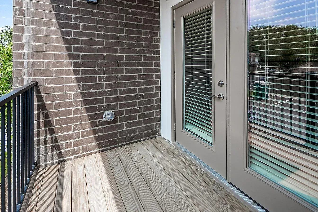 Balcony with wooden floor, brick wall, metal railing, and door with blinds.