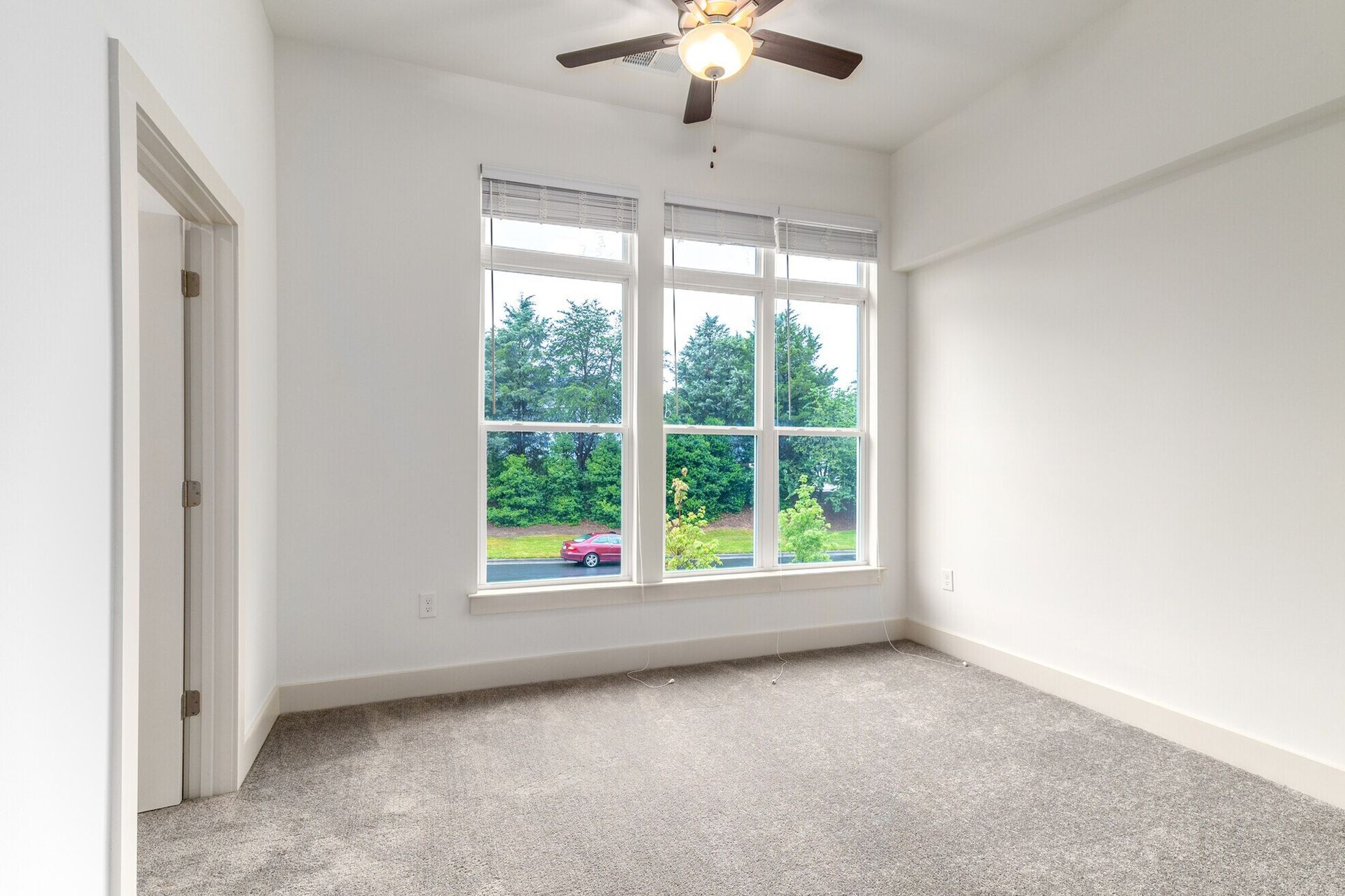 Empty white bedroom with large window, gray carpet, and a ceiling fan.