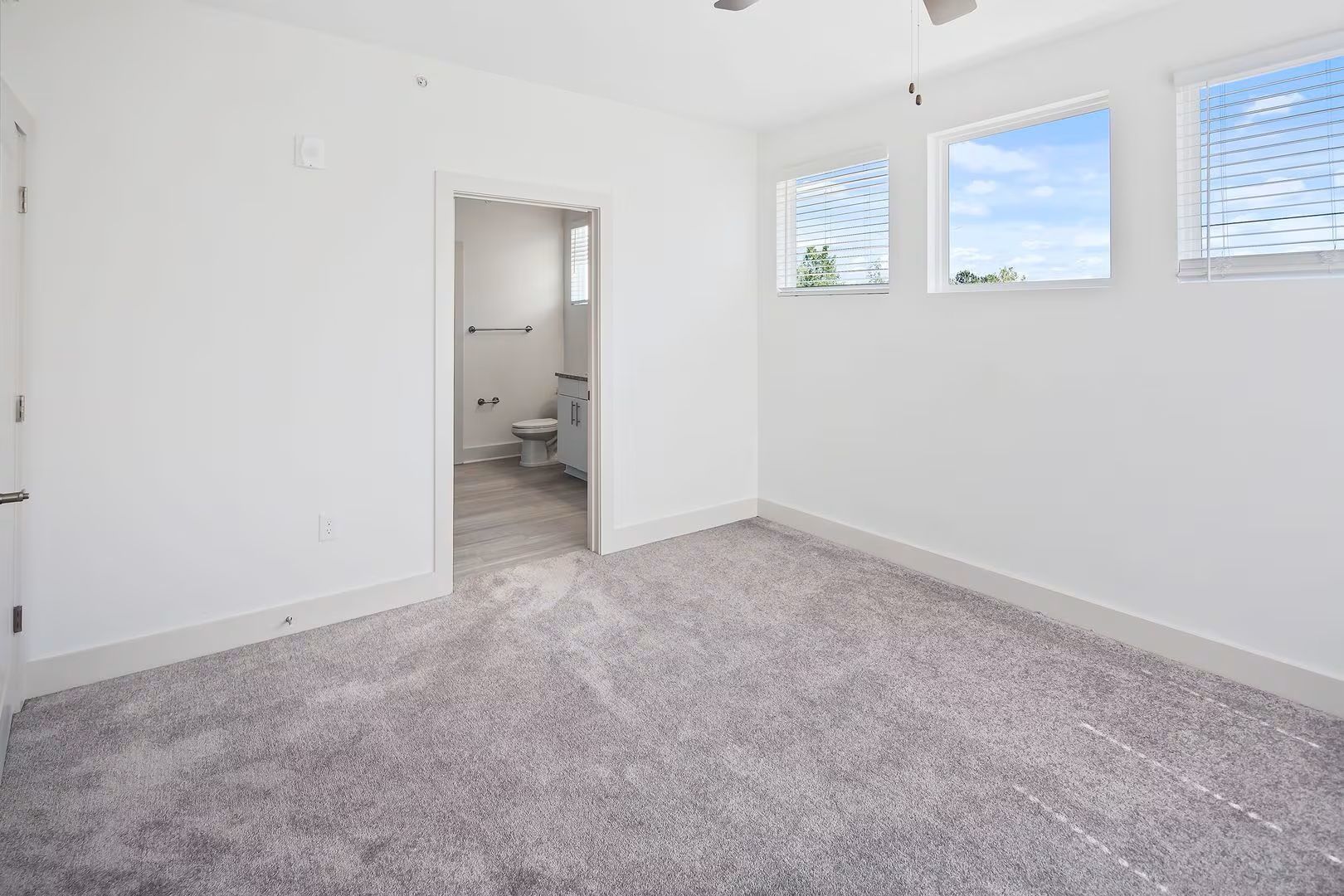 Empty white bedroom with gray carpet, connected bathroom, and three windows.