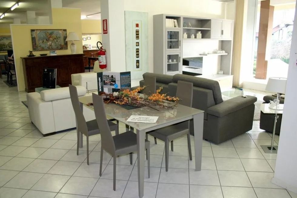 Showroom di mobili con un set da pranzo e due divani. Pavimento in piastrelle bianche, pareti neutre.