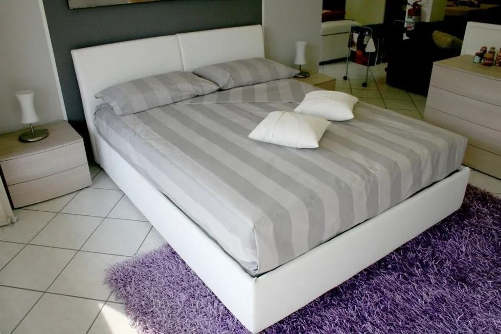 Letto imbottito bianco con lenzuola a righe, due cuscini e tappeto viola.