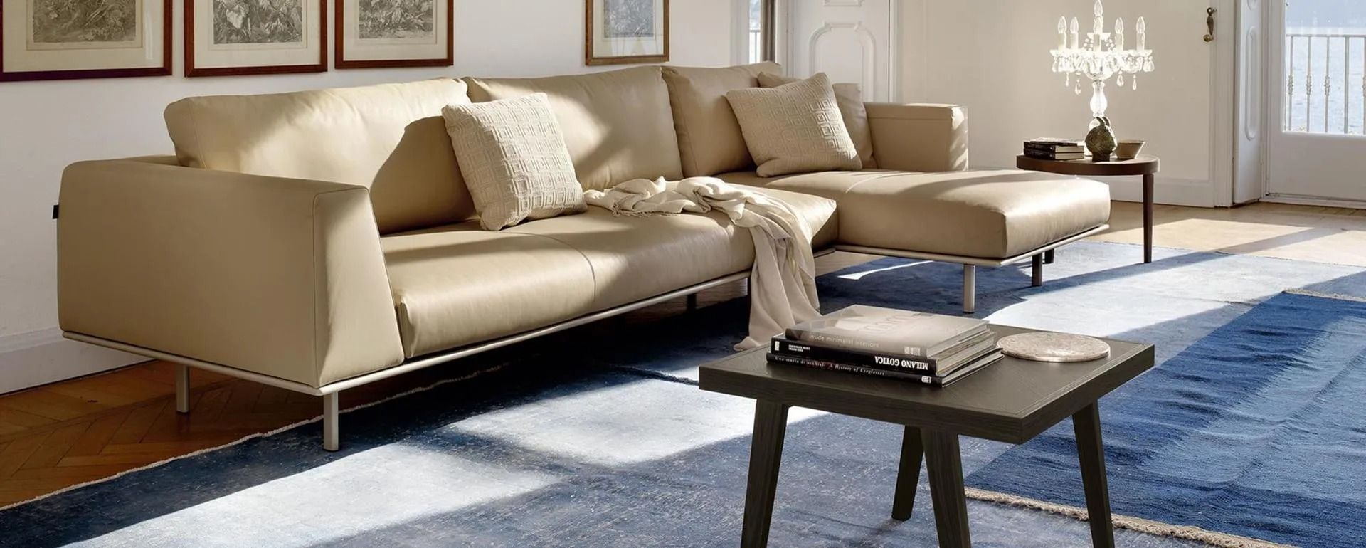Divano componibile beige su tappeto blu in una stanza luminosa con quadri incorniciati e un tavolino.