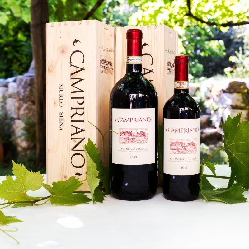 Due bottiglie di vino rosso accanto a una cassa di legno Campriano, con foglie di vite all'aperto.