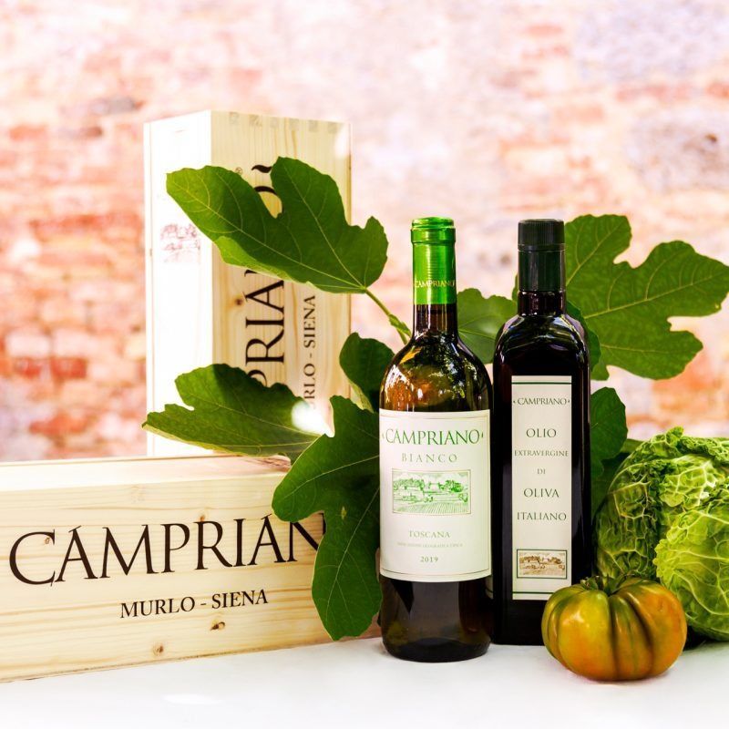 Bottiglie di olio d'oliva Campria, scatola, foglie e piccole zucche su uno sfondo rustico chiaro.