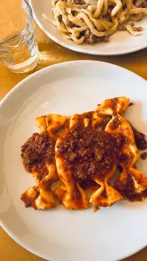 Piatto di pasta al ragù su un piatto bianco, con un altro piatto di pasta e un bicchiere d'acqua nelle vicinanze.