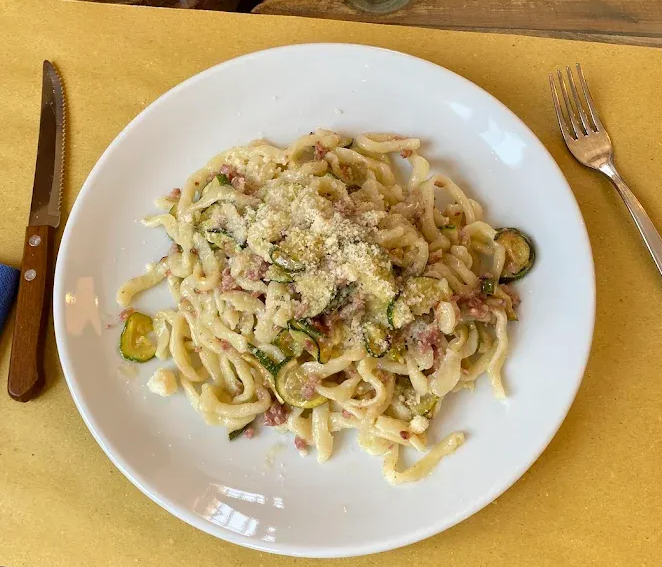 Piatto di pasta con formaggio grattugiato su un piatto bianco, con coltello e forchetta su una tovaglia gialla.
