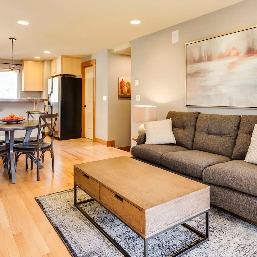 Living room with a brown couch, wooden coffee table, and a rug. Dining area visible in the background.