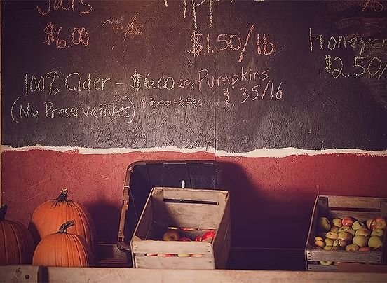 Blackboard with handwritten prices for apples, pumpkins, cider, and honey. Pumpkins and apples in crates below.