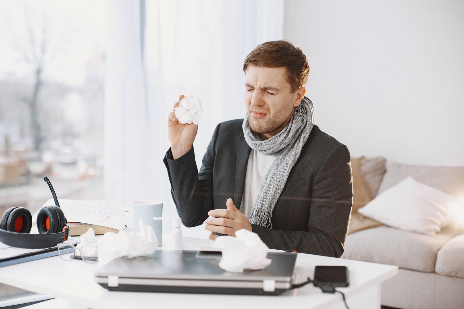 sick man in a grey scarf and blazer with a tissue in his hand making a face like he just sneezed