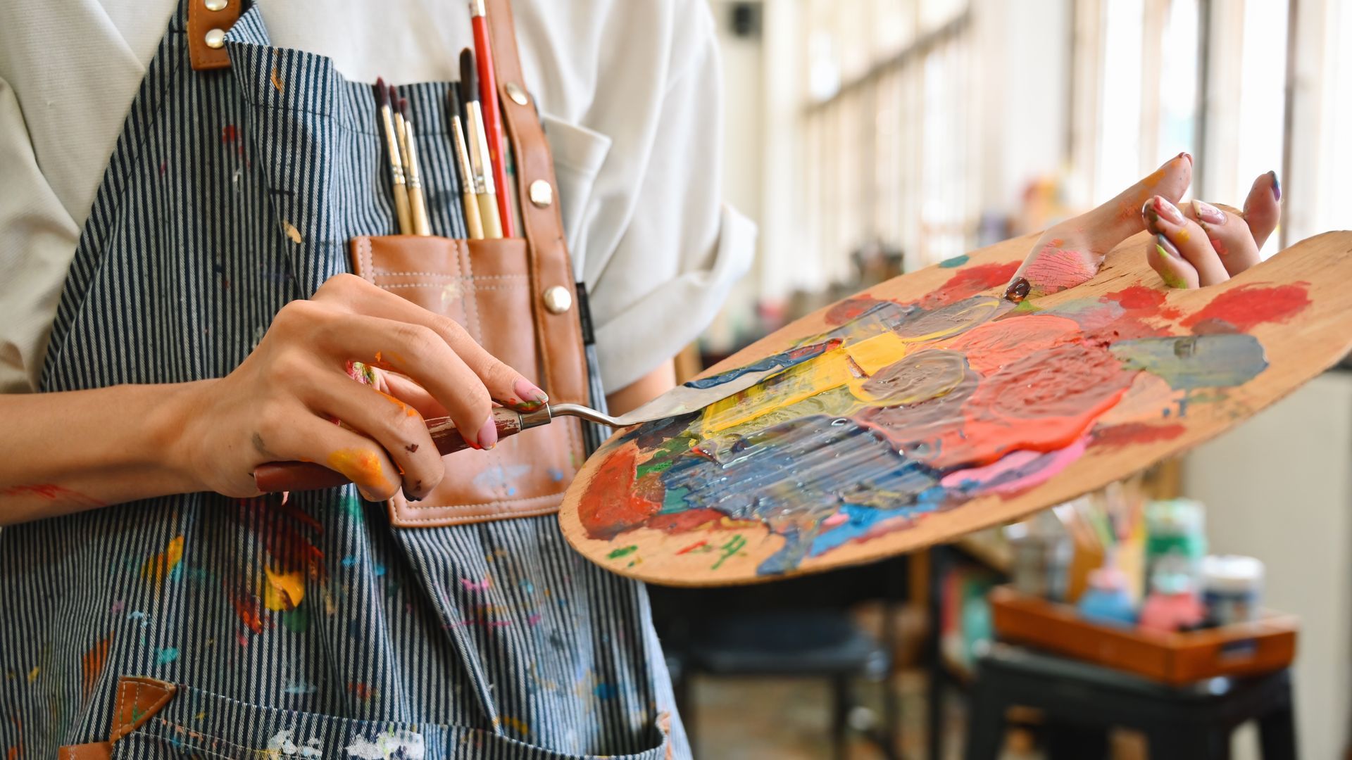 Artist using a palette knife to mix paint on a wooden palette, wearing an apron, in a studio.