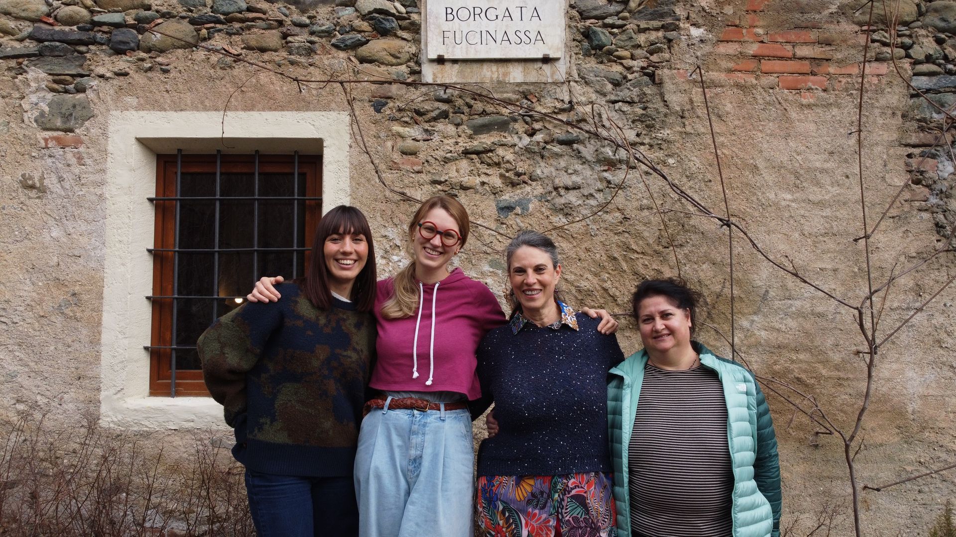 Quattro persone sono in piedi, sorridenti, davanti a un edificio in pietra con una piccola finestra con le sbarre e una targa commemorativa.