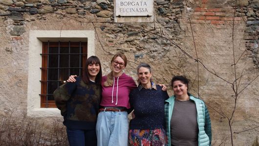 Quattro persone sono in piedi, sorridenti, davanti a un edificio in pietra con una piccola finestra con le sbarre e una targa commemorativa.