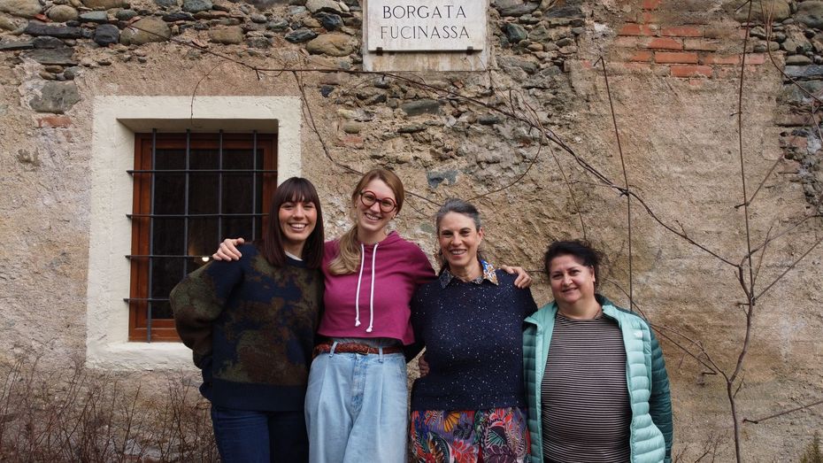 Quattro persone sono in piedi, sorridenti, davanti a un edificio in pietra con una piccola finestra con le sbarre e una targa commemorativa.