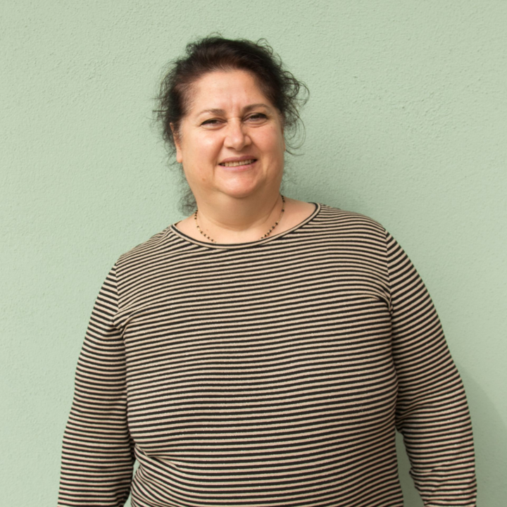 Persona sorridente con indosso una camicia a maniche lunghe a righe marrone scuro e chiaro, in piedi davanti a un muro verde chiaro.