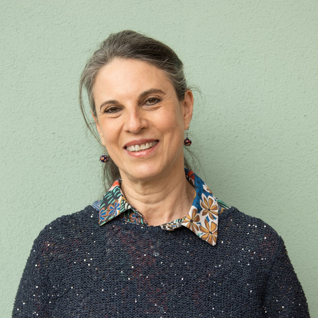 Una persona sorridente su uno sfondo verde chiaro, con indosso un maglione blu scuro a trama grossa sopra una camicia con colletto a fiori.