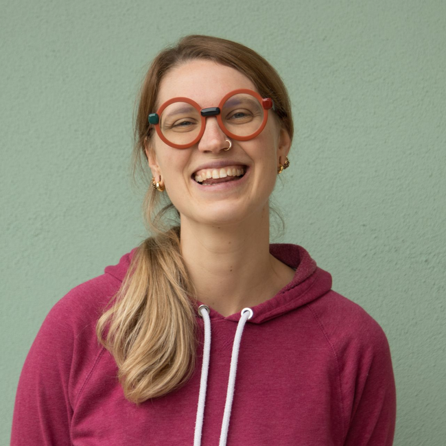 Una persona sorridente con occhiali rotondi dalla montatura arancione e una felpa con cappuccio color magenta, sullo sfondo di un muro verde chiaro.