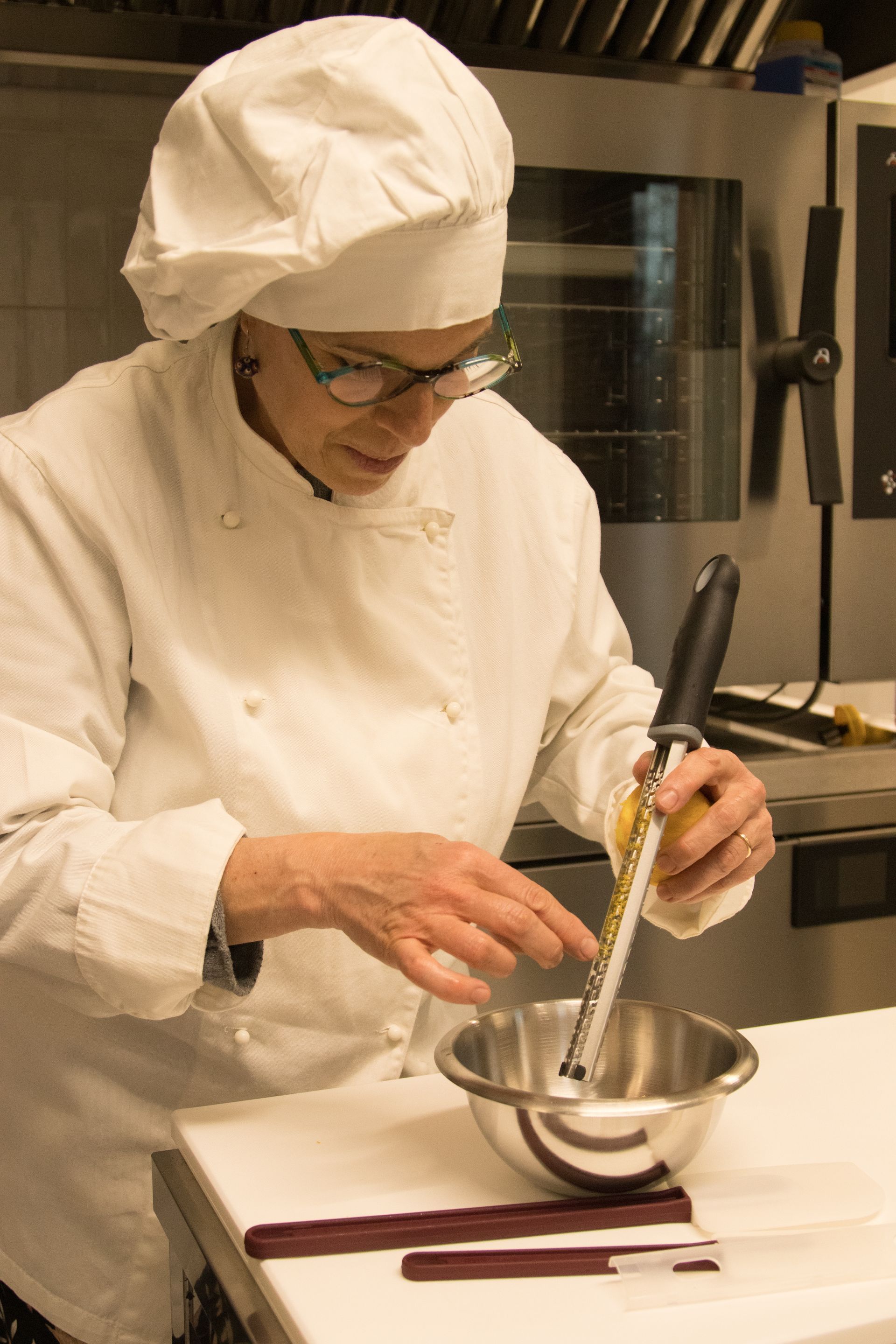 In cucina, uno chef in uniforme e cappello bianchi utilizza una grattugia per agrumi sopra una ciotola di metallo.