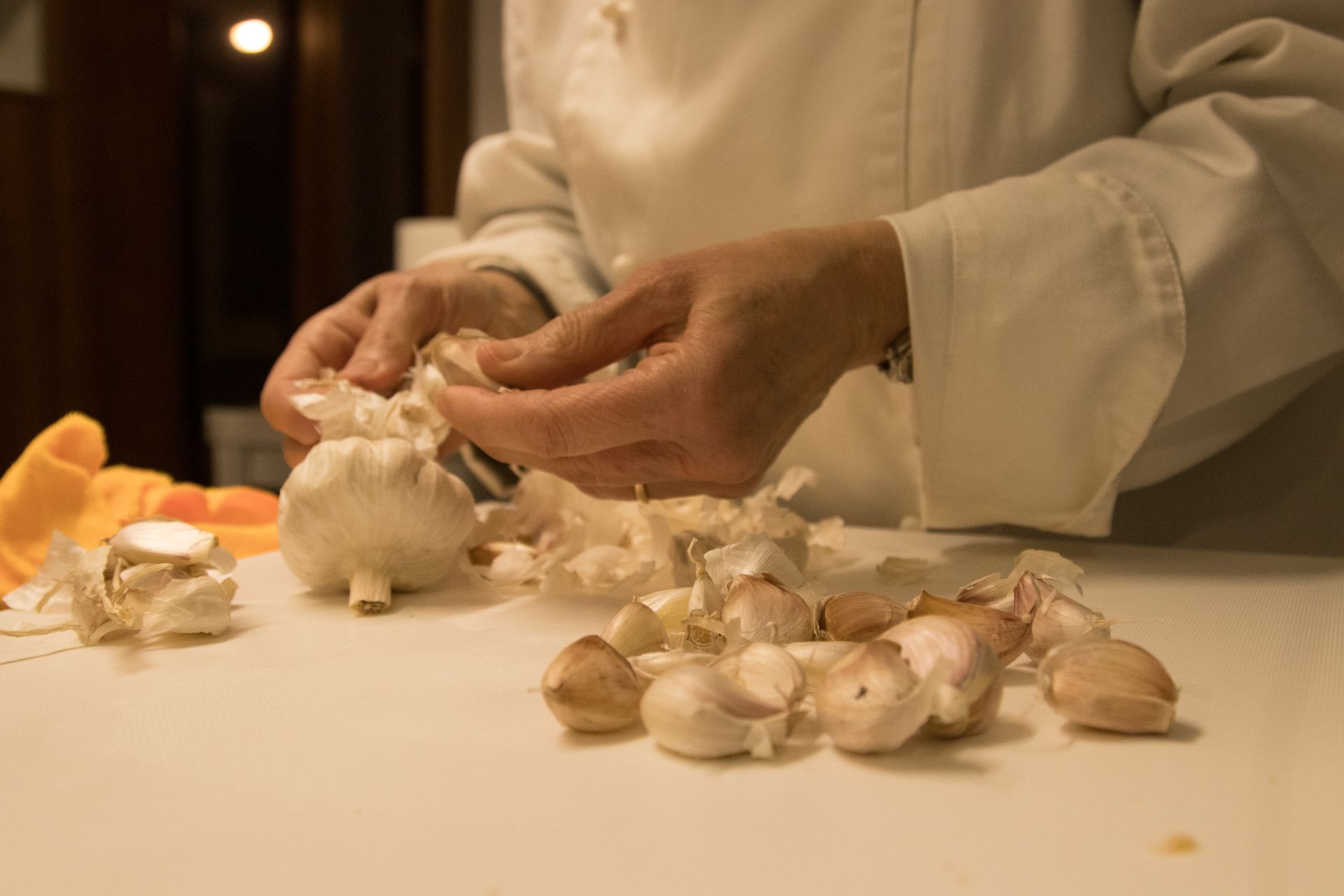 Uno chef in uniforme bianca sbuccia singoli spicchi d'aglio da un bulbo e li deposita su un tagliere bianco.