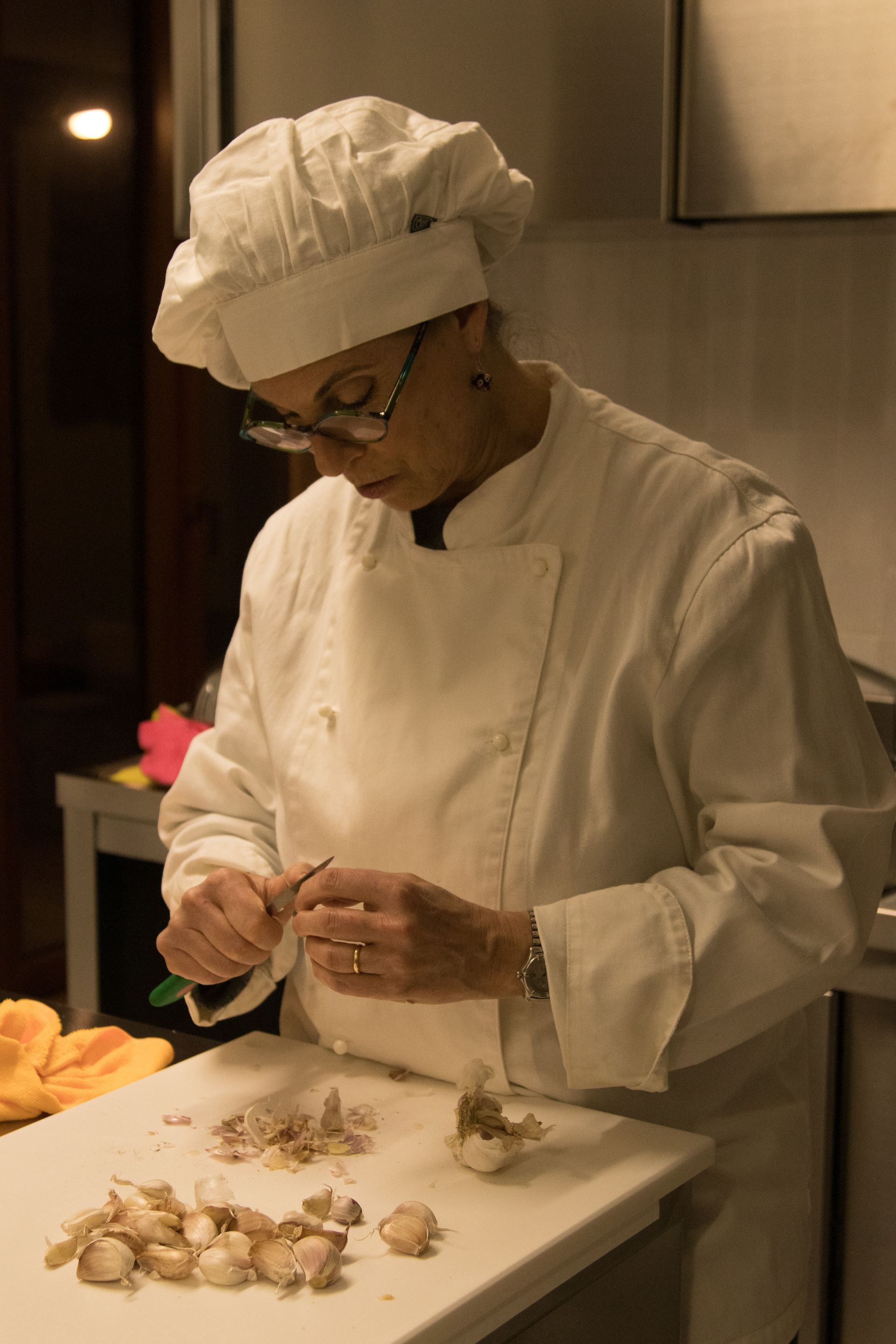 In una cucina, uno chef in uniforme e cappello bianchi sbuccia spicchi d'aglio su un tagliere bianco.