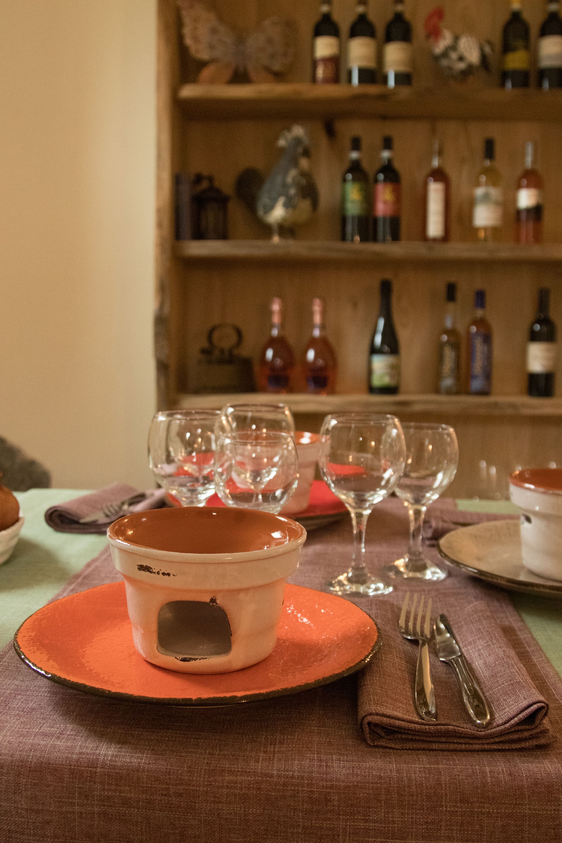 Una tavola apparecchiata con un piatto arancione e uno scaldavivande in terracotta, bicchieri da vino, posate e, sullo sfondo, una rastrelliera per il vino.