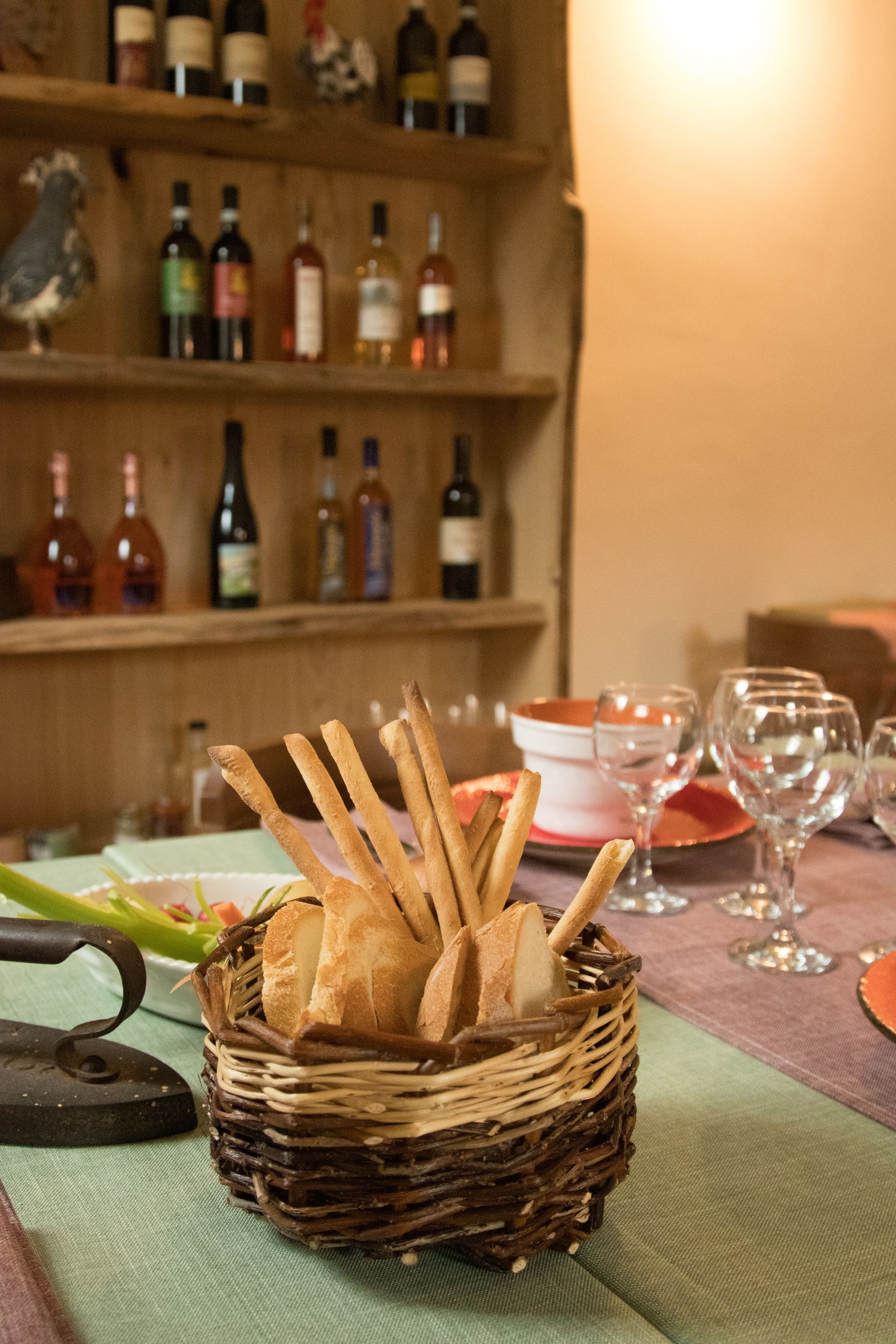 Su un tavolo apparecchiato per la cena, un cesto di vimini contenente grissini e fette di pane è appoggiato, mentre sullo sfondo si vede uno scaffale pieno di bottiglie di vino.