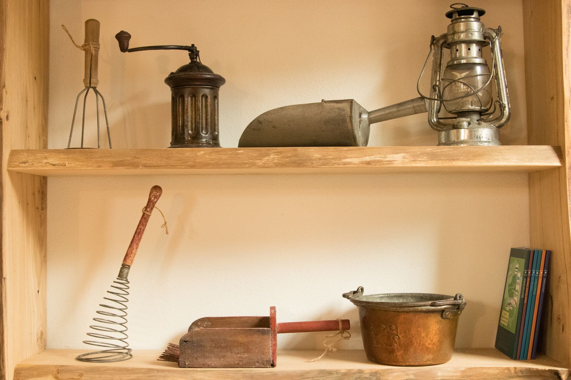Due mensole in legno espongono vari utensili da cucina d'epoca, tra cui un macinino, una lanterna, una frusta e una pentola di rame.