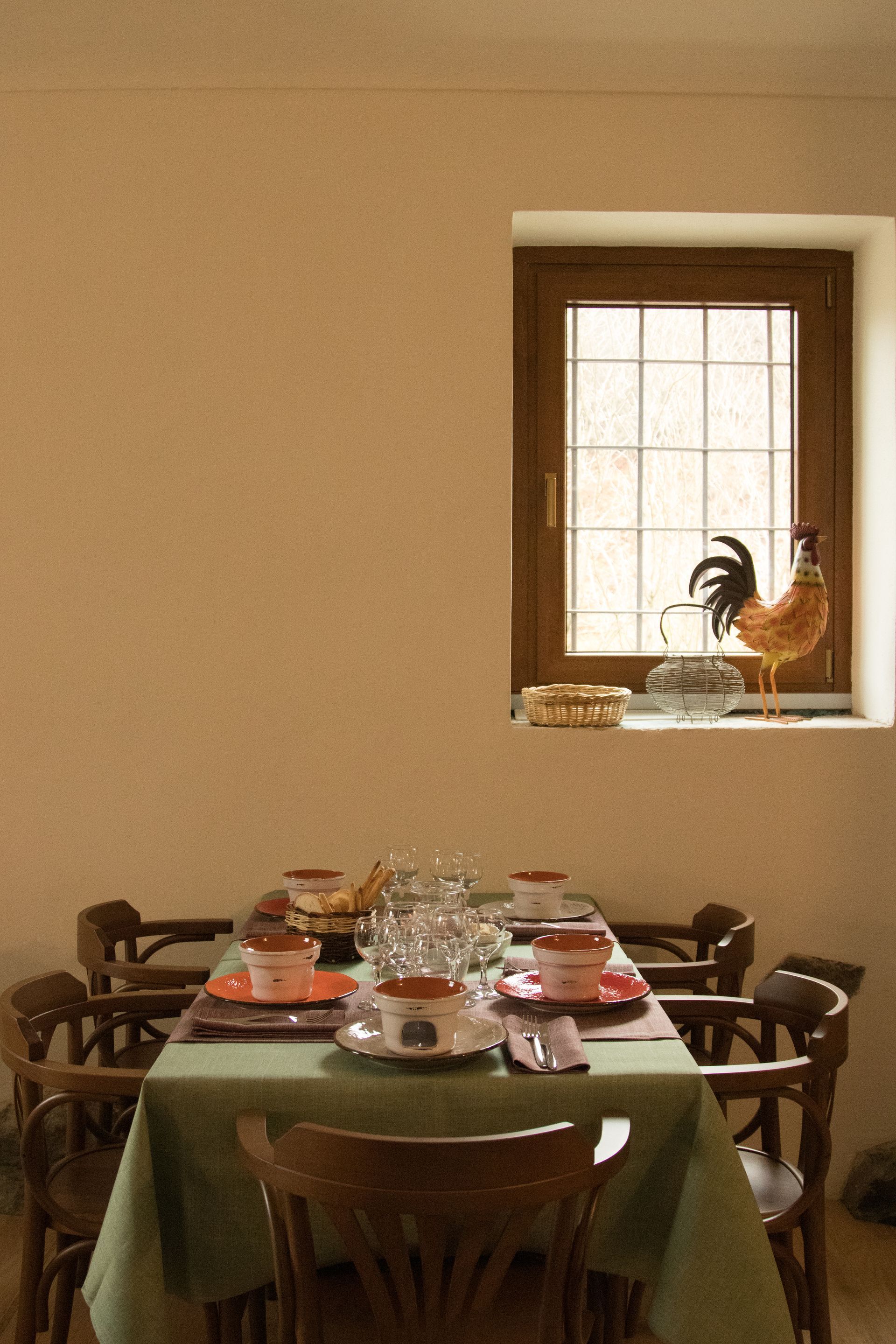 Un tavolo da pranzo per quattro persone con tovaglia verde chiaro e sedie di legno, posizionato davanti a una finestra con una statuetta di un gallo.