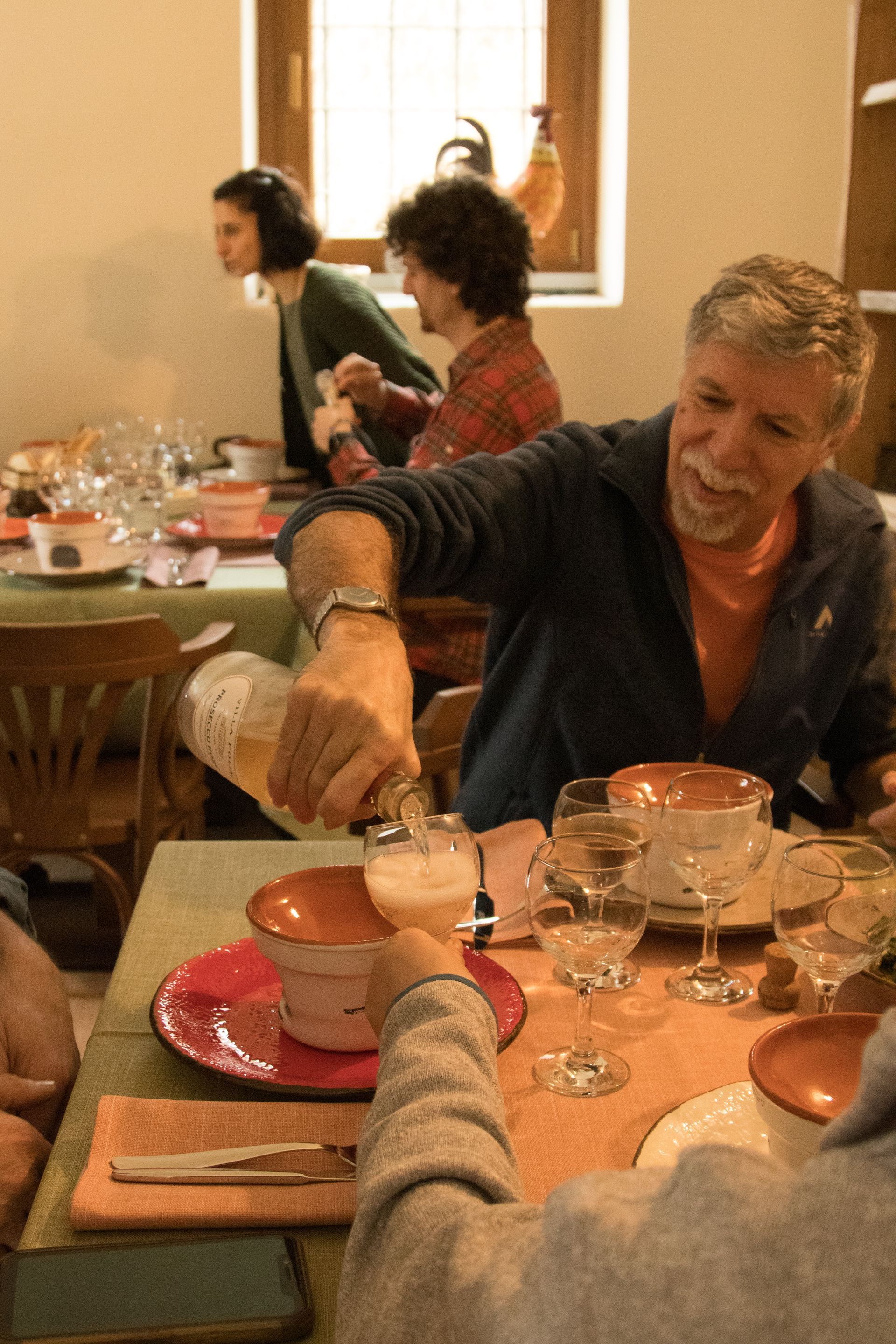 Una persona sorridente versa del vino bianco in un bicchiere a tavola, mentre altri commensali cenano sullo sfondo.