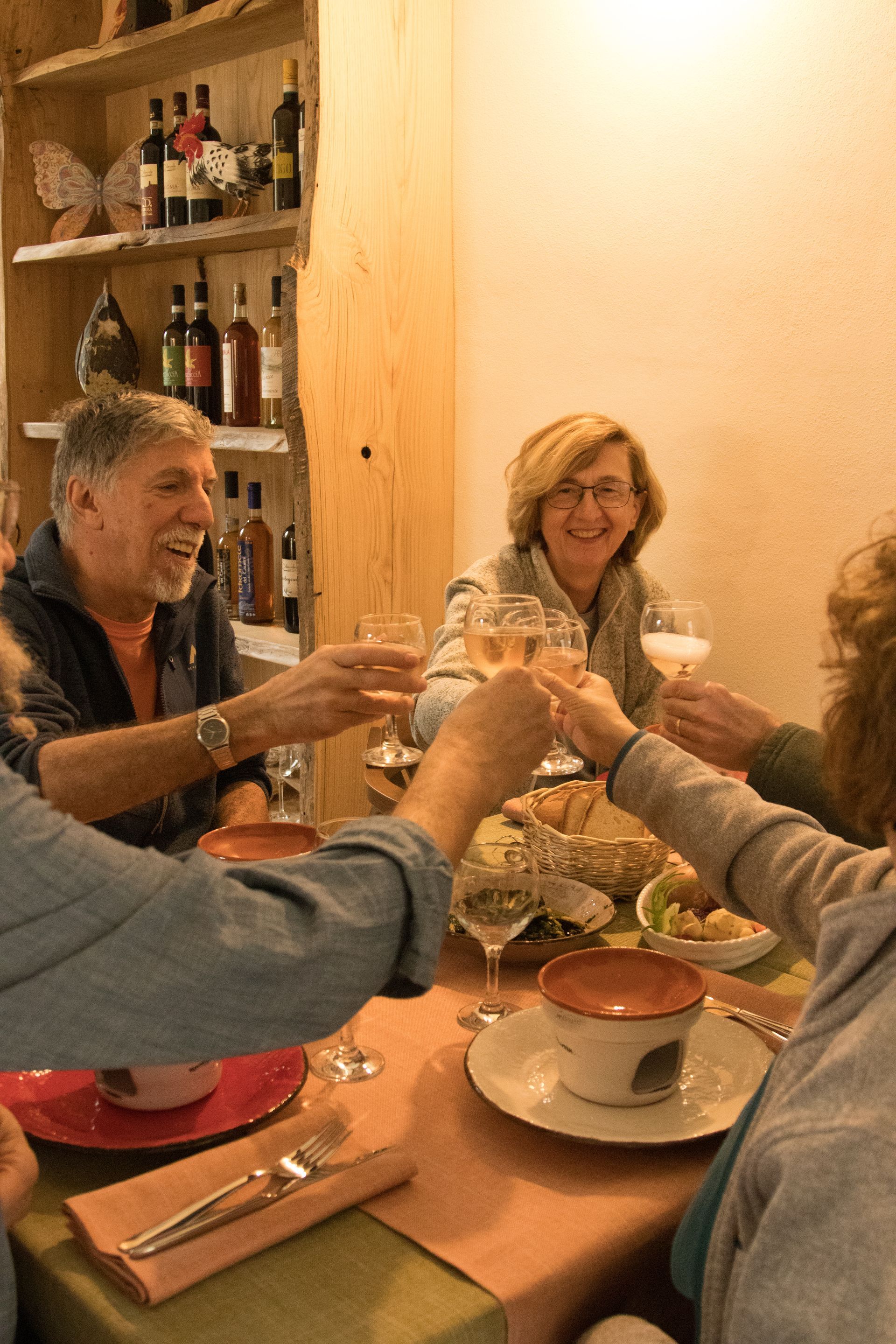 Durante una cena in un accogliente ristorante, alcune persone brindano con i calici di vino, con uno scaffale di legno pieno di bottiglie sullo sfondo.